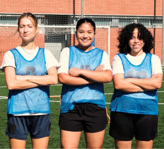 Three teammates in blue practice jerseys on a soccer field